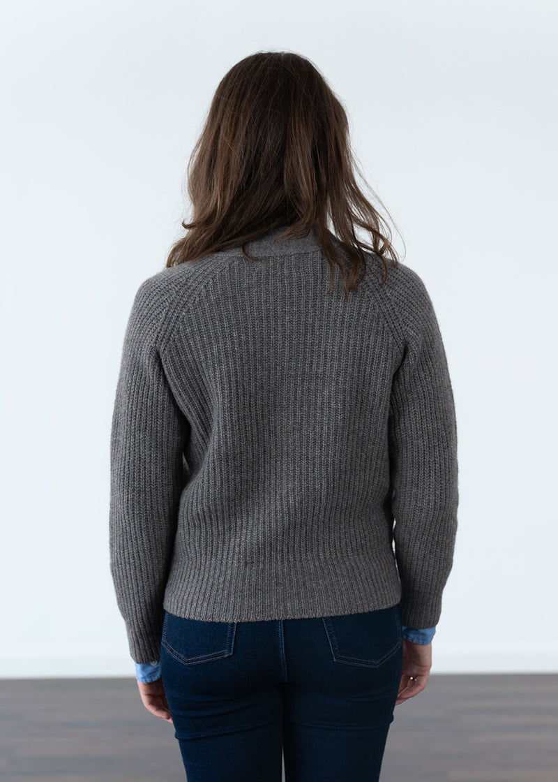 A woman viewed from behind wearing a brown ribbed knit cardigan and dark blue jeans, standing against a plain white wall.
