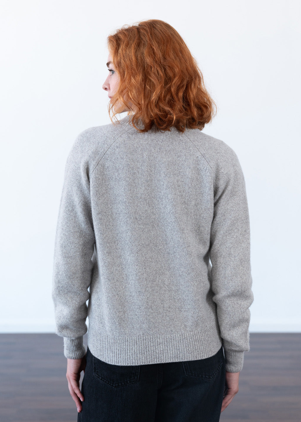 A woman with wavy red hair is shown from behind wearing a light gray knit sweater and dark jeans, standing against a white wall with a dark wood floor.