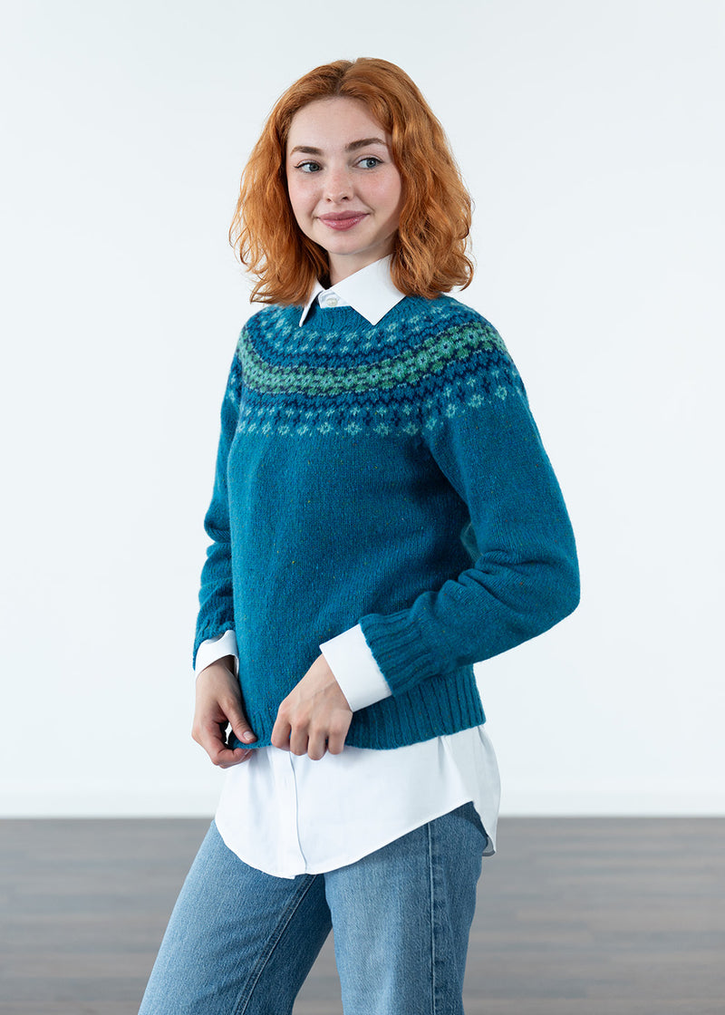 A woman with red hair wearing a teal blue Fair Isle knit sweater over a white collared shirt and blue jeans, standing against a clean white studio background.