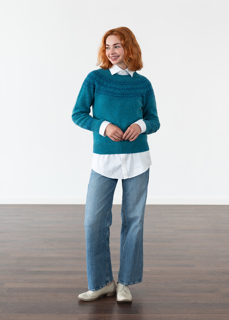 A woman stands in a studio wearing a teal fair isle jumper over a white collared shirt, blue jeans, and white shoes, against a plain white wall and dark wood floor.