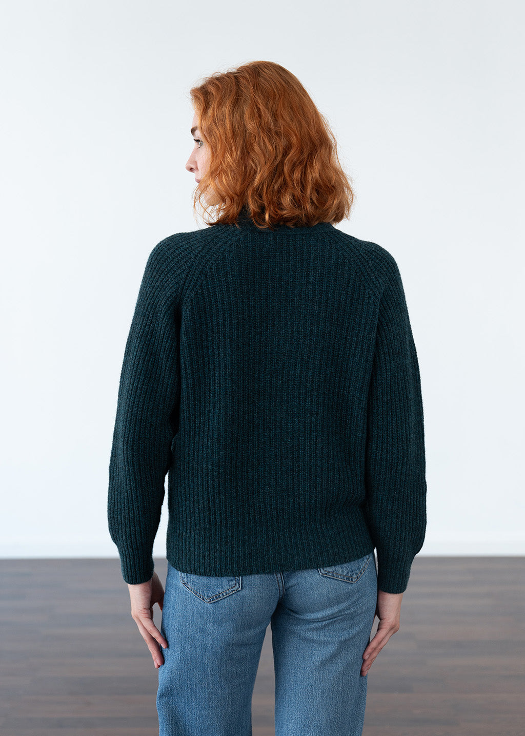A woman with red hair is shown from the back wearing a dark teal ribbed knit cardigan and blue jeans against a white studio background.