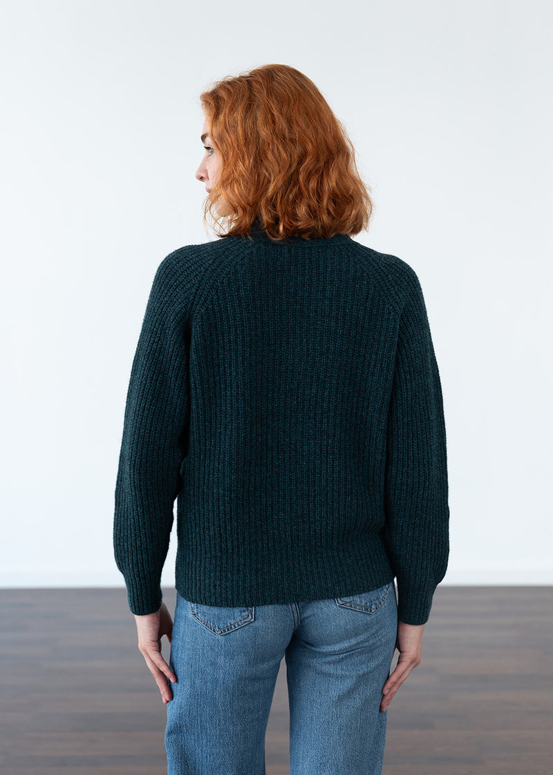 A woman with red hair is shown from the back wearing a dark teal ribbed knit cardigan and blue jeans against a white studio background.