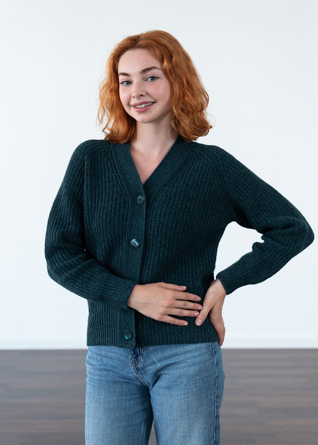 A woman with red hair wearing a dark teal ribbed knit cardigan and blue jeans, standing against a clean white studio background.