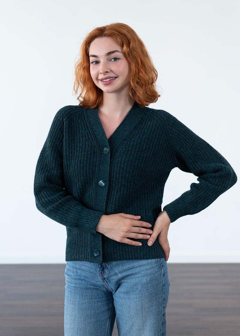 A woman with red hair wearing a dark teal ribbed knit cardigan and blue jeans, standing against a clean white studio background.