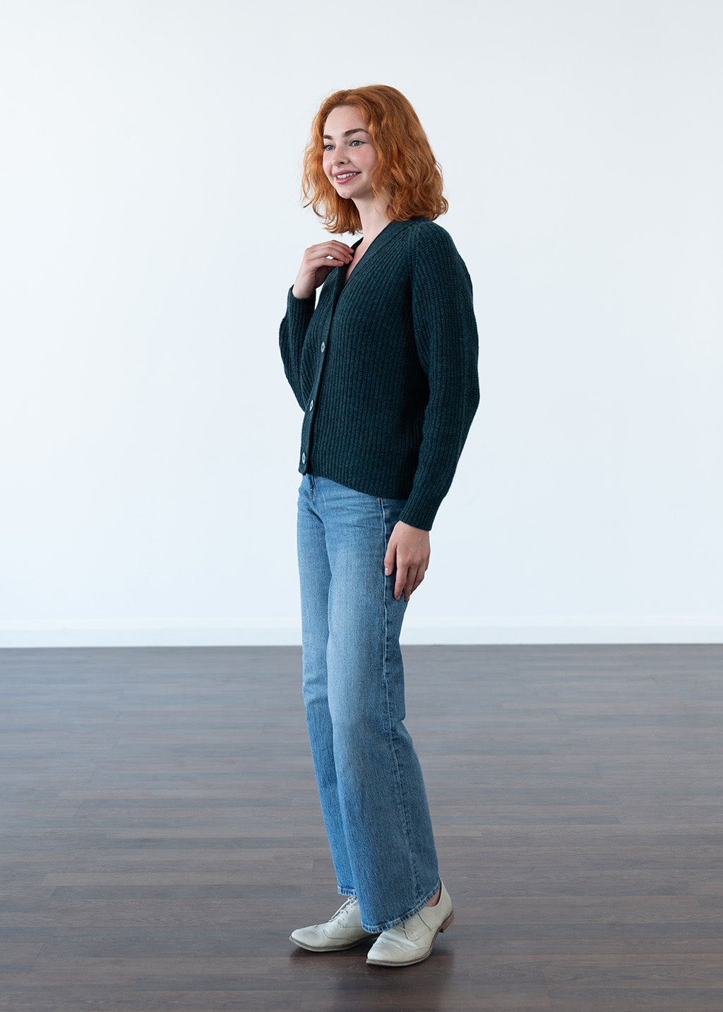 A woman with red hair stands in a studio wearing a dark teal ribbed cardigan, light blue jeans, and white shoes against a white wall and dark wood floor.