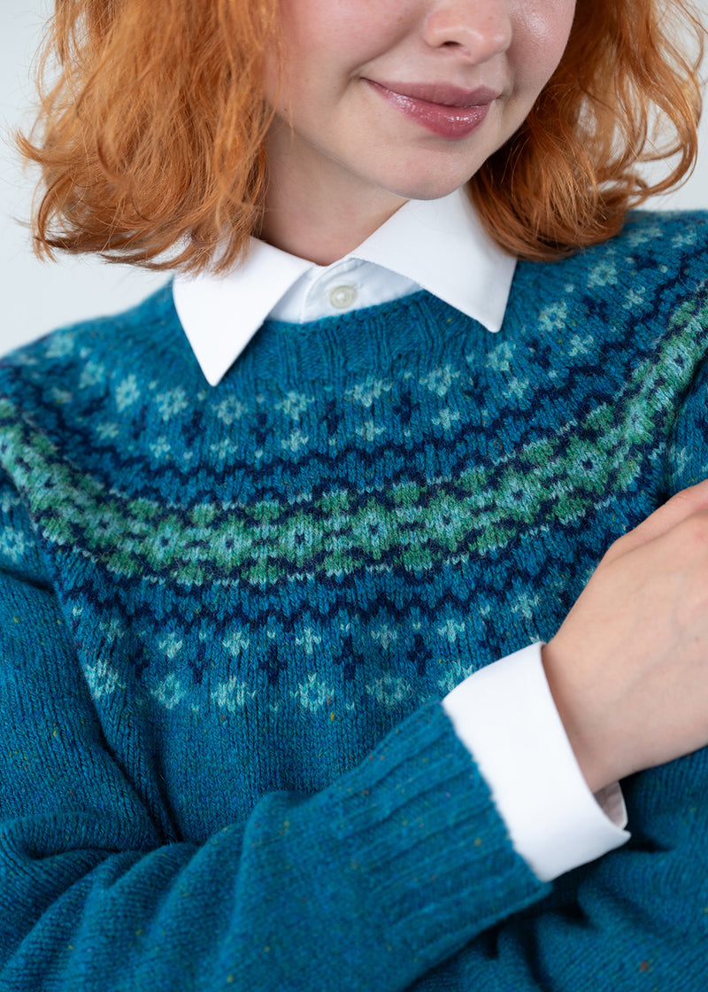 A close-up of a person wearing a teal and green patterned knit sweater over a white collared shirt. The sweater features a Fair Isle style pattern with shades of blue, green, and navy.