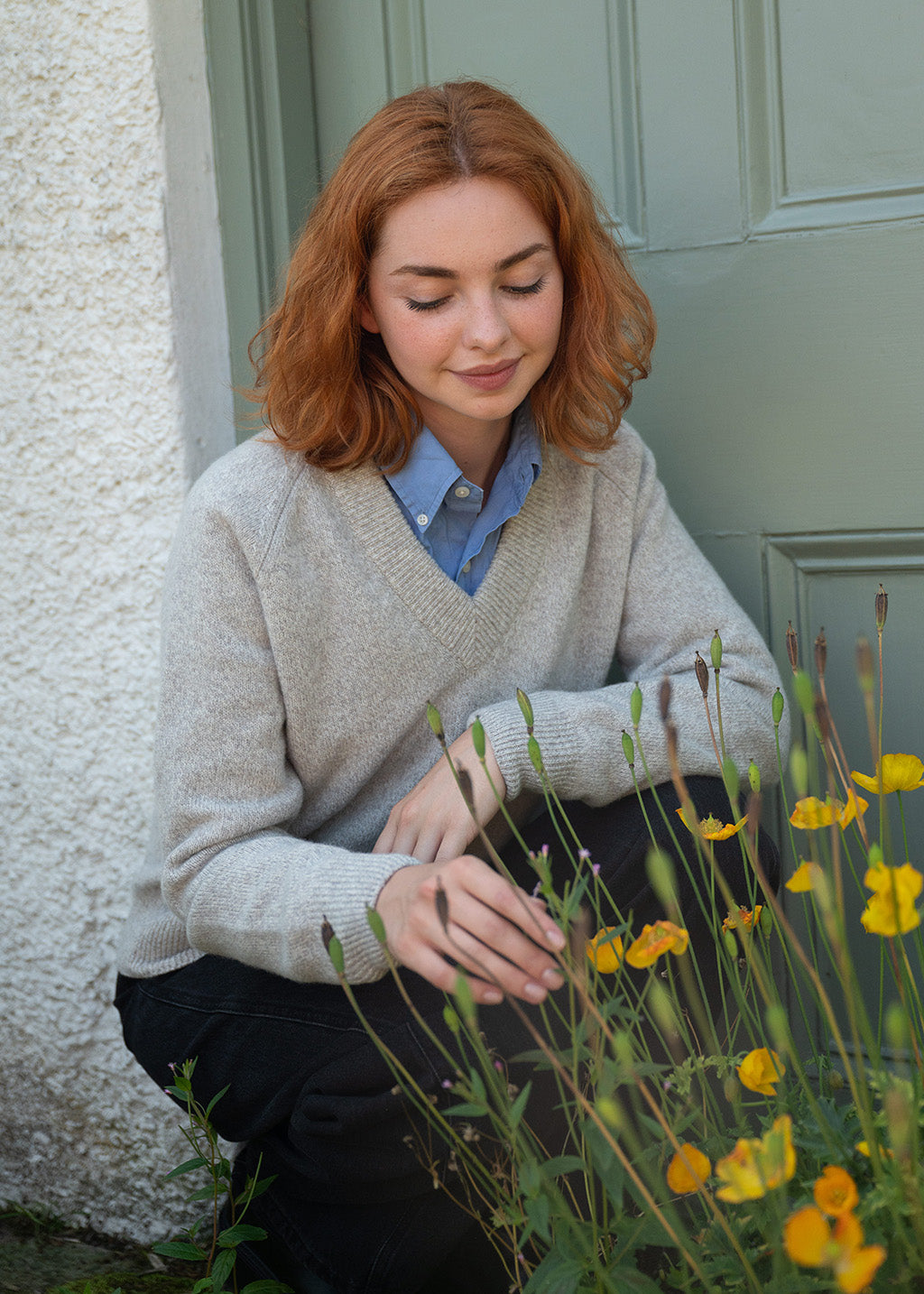 A woman with red hair crouches outdoors in front of a sage green door, wearing a grey marl V-neck sweater over a blue collared shirt and dark pants,jeans with yellow poppies in the foreground.